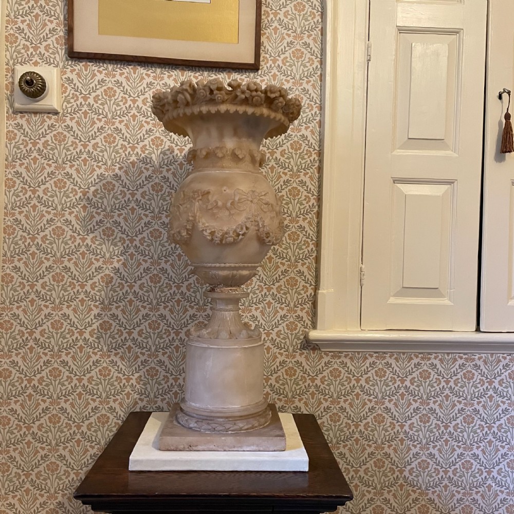 A carved 19th-century cream alabaster urn featuring floral rim detailing, draped garland festoons, and a tall cylindrical pedestal base. View II