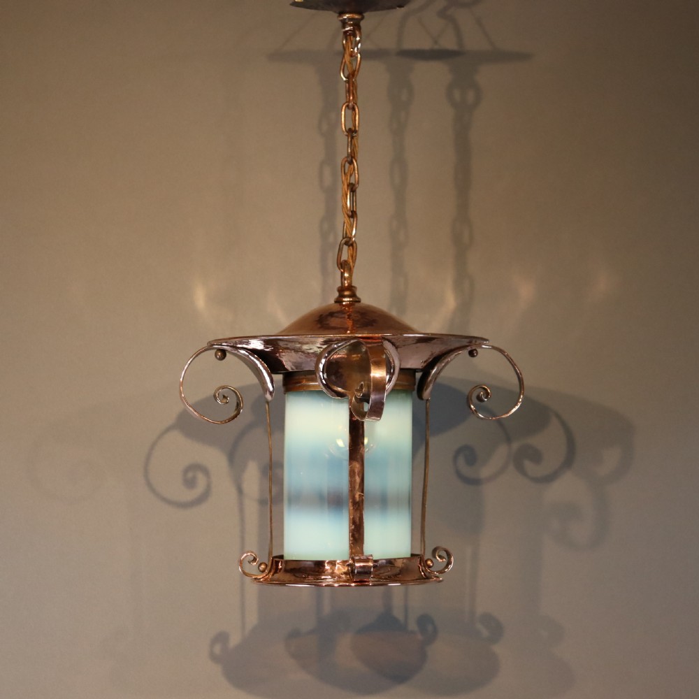 An Arts and Crafts copper pendant light featuring a cylindrical opalescent glass shade held within a copper frame with scrolled side accents and a suspension chain.