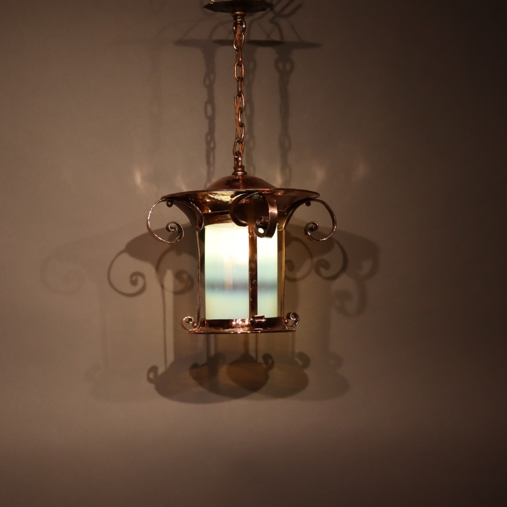 An Arts and Crafts copper pendant light featuring a cylindrical opalescent glass shade held within a copper frame with scrolled side accents and a suspension chain. View II