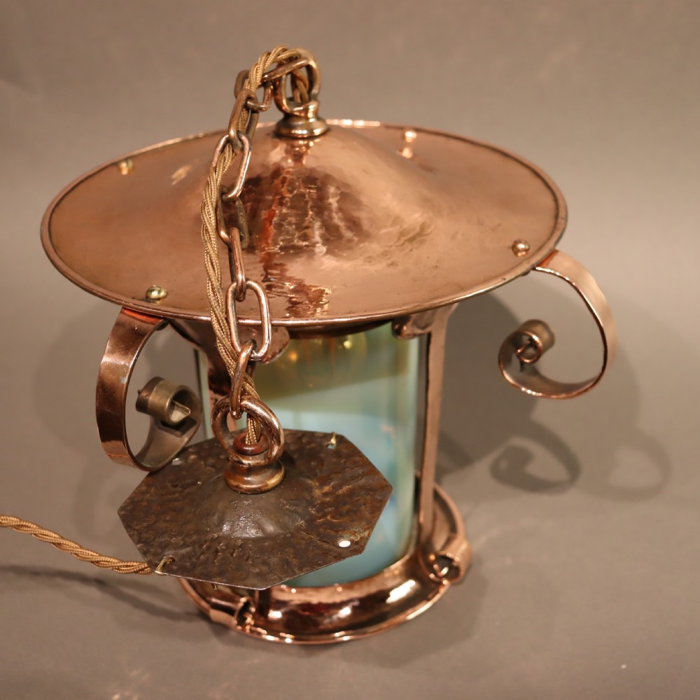 An Arts and Crafts copper pendant light featuring a cylindrical opalescent glass shade held within a copper frame with scrolled side accents and a suspension chain. View III