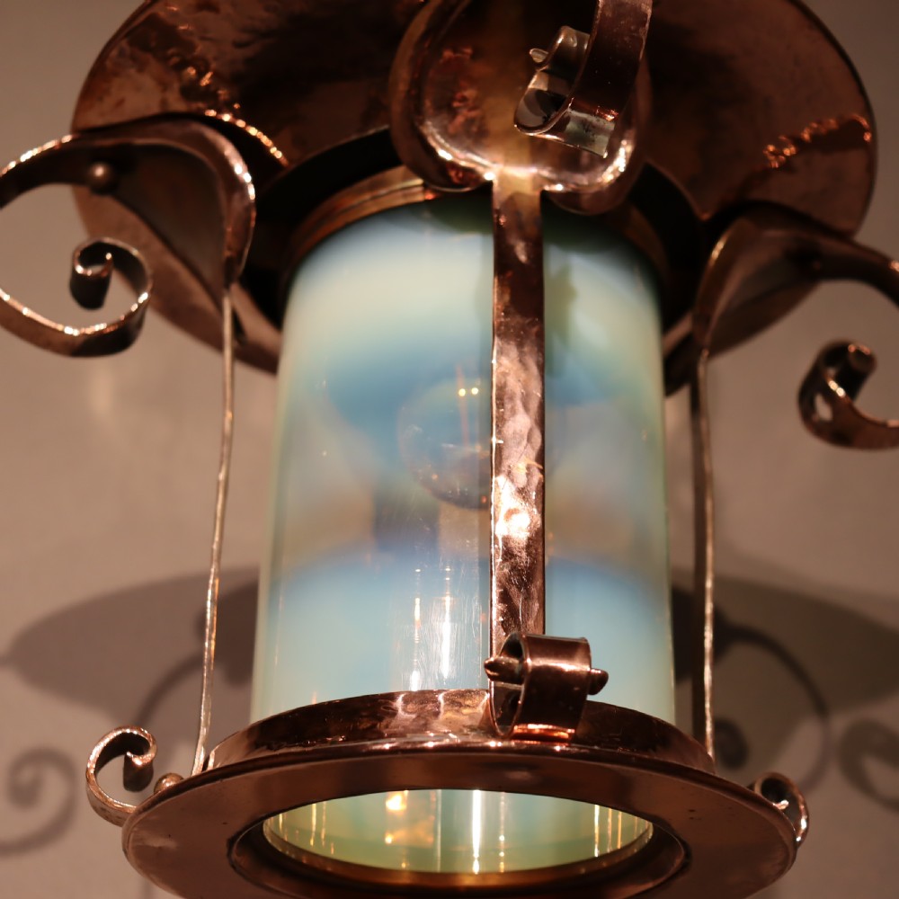 An Arts and Crafts copper pendant light featuring a cylindrical opalescent glass shade held within a copper frame with scrolled side accents and a suspension chain. View IV