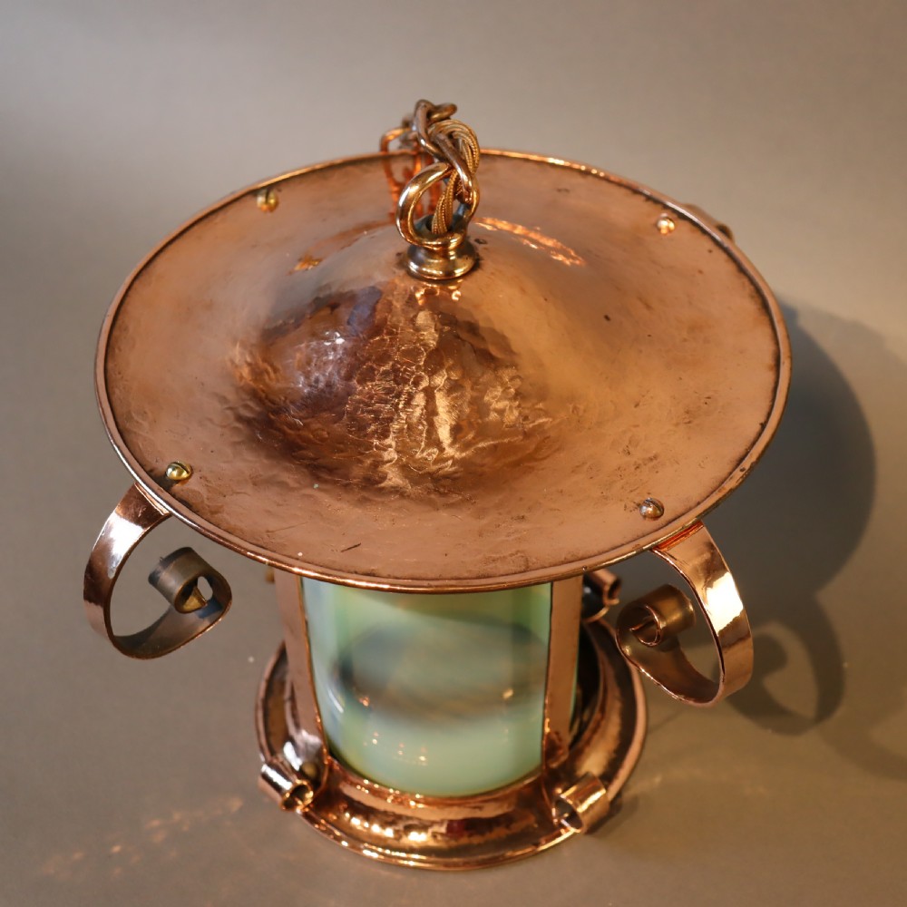 An Arts and Crafts copper pendant light featuring a cylindrical opalescent glass shade held within a copper frame with scrolled side accents and a suspension chain. View V