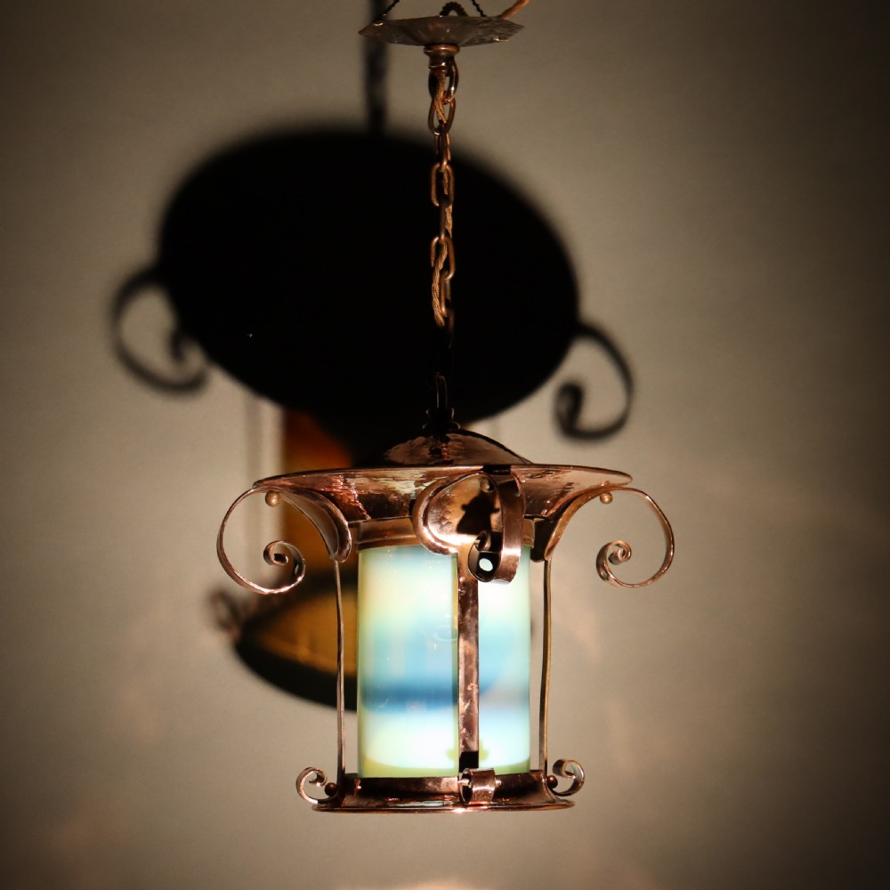 An Arts and Crafts copper pendant light featuring a cylindrical opalescent glass shade held within a copper frame with scrolled side accents and a suspension chain. View VI
