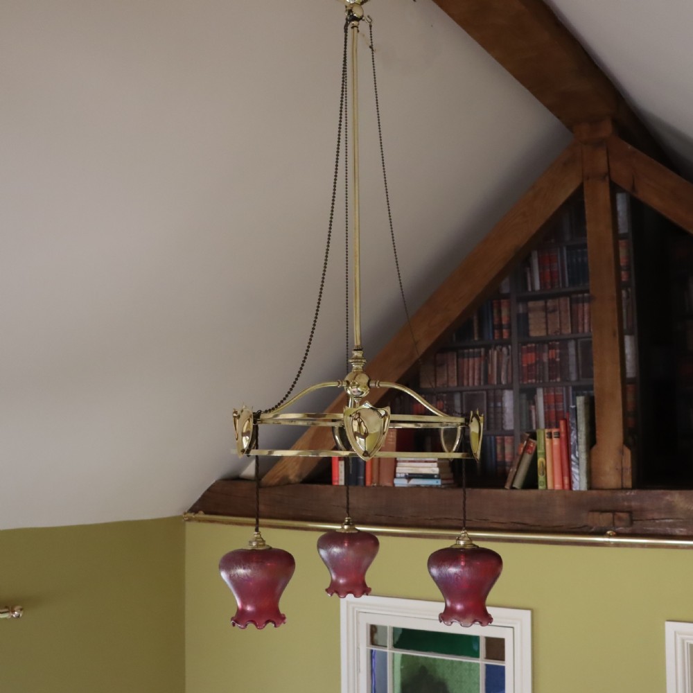 An antique Arts and Crafts brass three-branch ceiling light featuring a central stem, chain suspension, a circular brass ring with stylized motifs, and three hanging red cranberry glass shades.