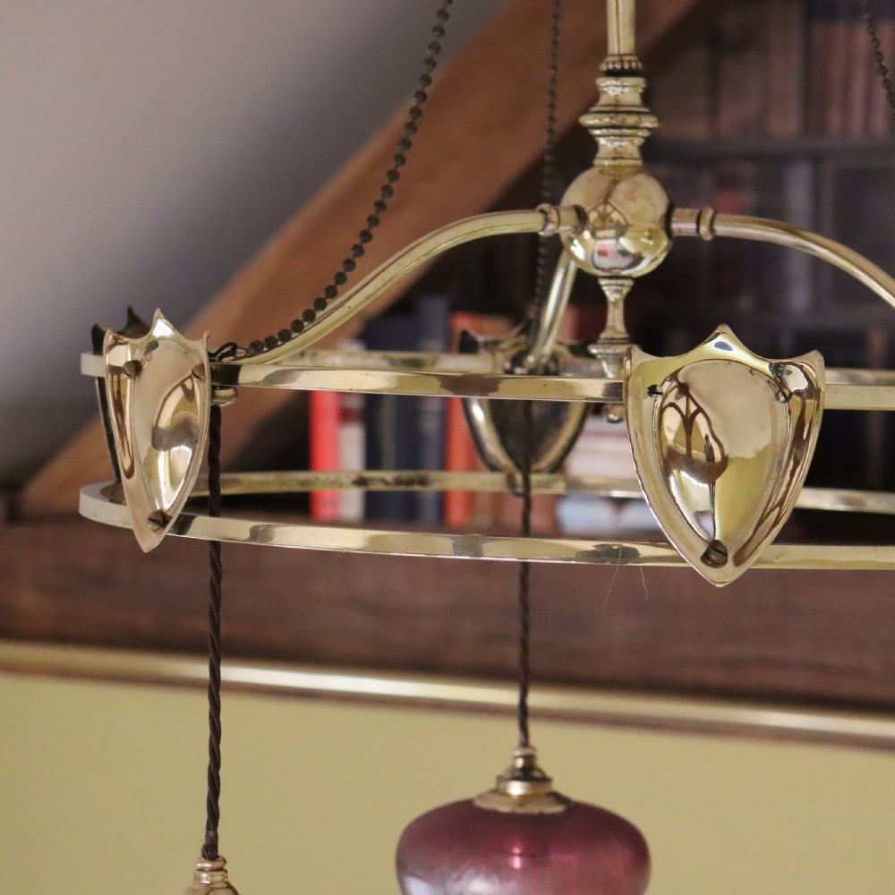 An antique Arts and Crafts brass three-branch ceiling light featuring a central stem, chain suspension, a circular brass ring with stylized motifs, and three hanging red cranberry glass shades. View II