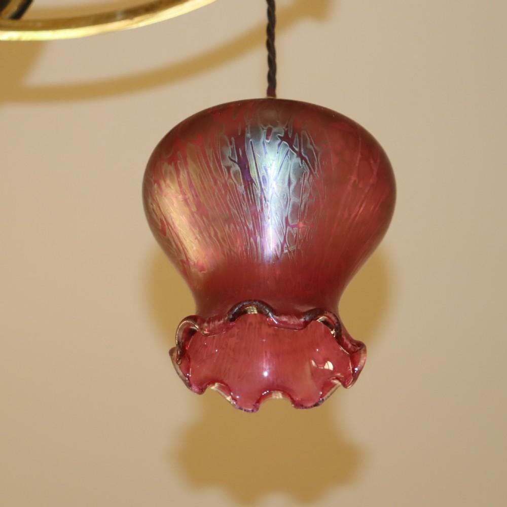 An antique Arts and Crafts brass three-branch ceiling light featuring a central stem, chain suspension, a circular brass ring with stylized motifs, and three hanging red cranberry glass shades. View III