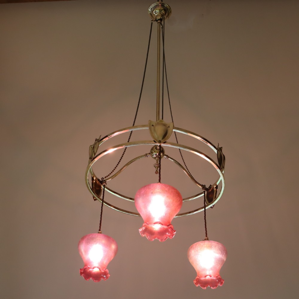 An antique Arts and Crafts brass three-branch ceiling light featuring a central stem, chain suspension, a circular brass ring with stylized motifs, and three hanging red cranberry glass shades. View IV