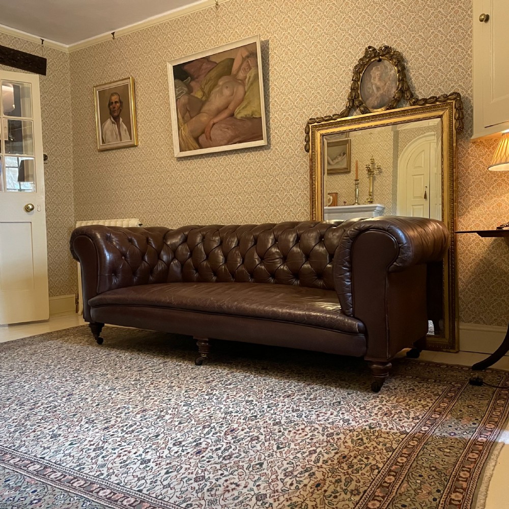 A three-seater brown leather Chesterfield sofa featuring deep button tufting, scrolled arms, and turned wooden legs with castors.