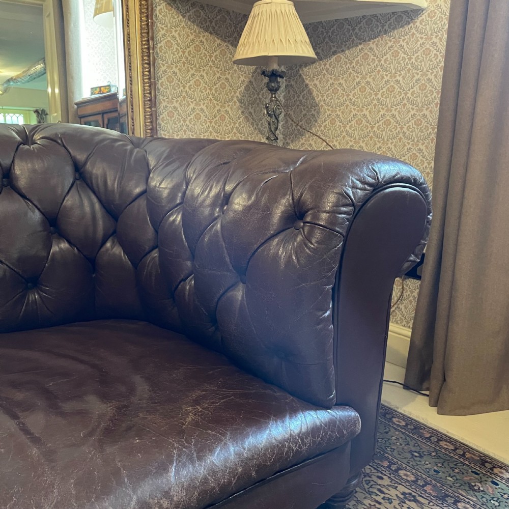 A three-seater brown leather Chesterfield sofa featuring deep button tufting, scrolled arms, and turned wooden legs with castors. View IV