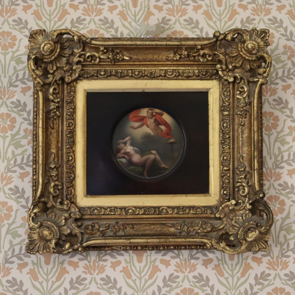 A small circular early 19th-century mythological oil painting depicting a reclining nude nymph and floating male god, encased in a square, highly ornate giltwood frame.
