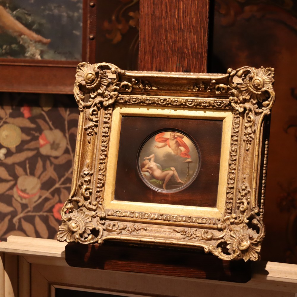 A small circular early 19th-century mythological oil painting depicting a reclining nude nymph and floating male god, encased in a square, highly ornate giltwood frame. View X