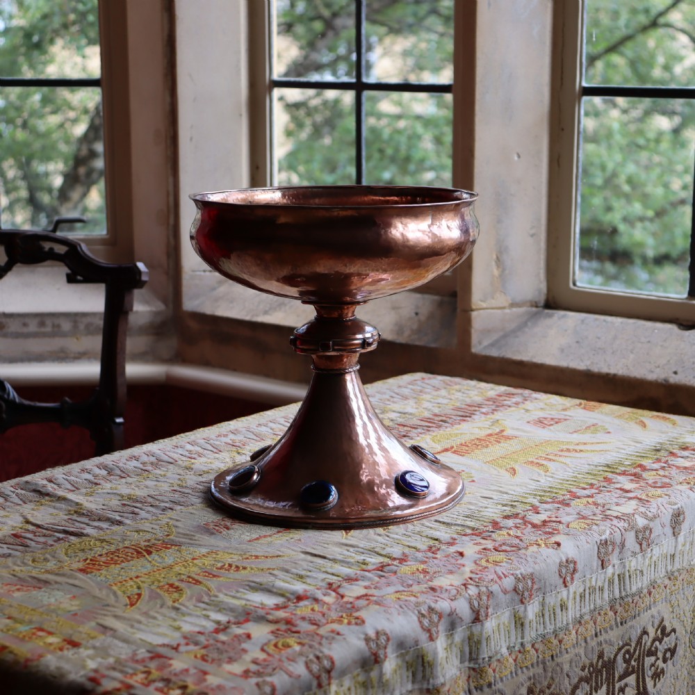 A hand-hammered copper pedestal bowl featuring a flared base set with circular blue enamel cabochons.