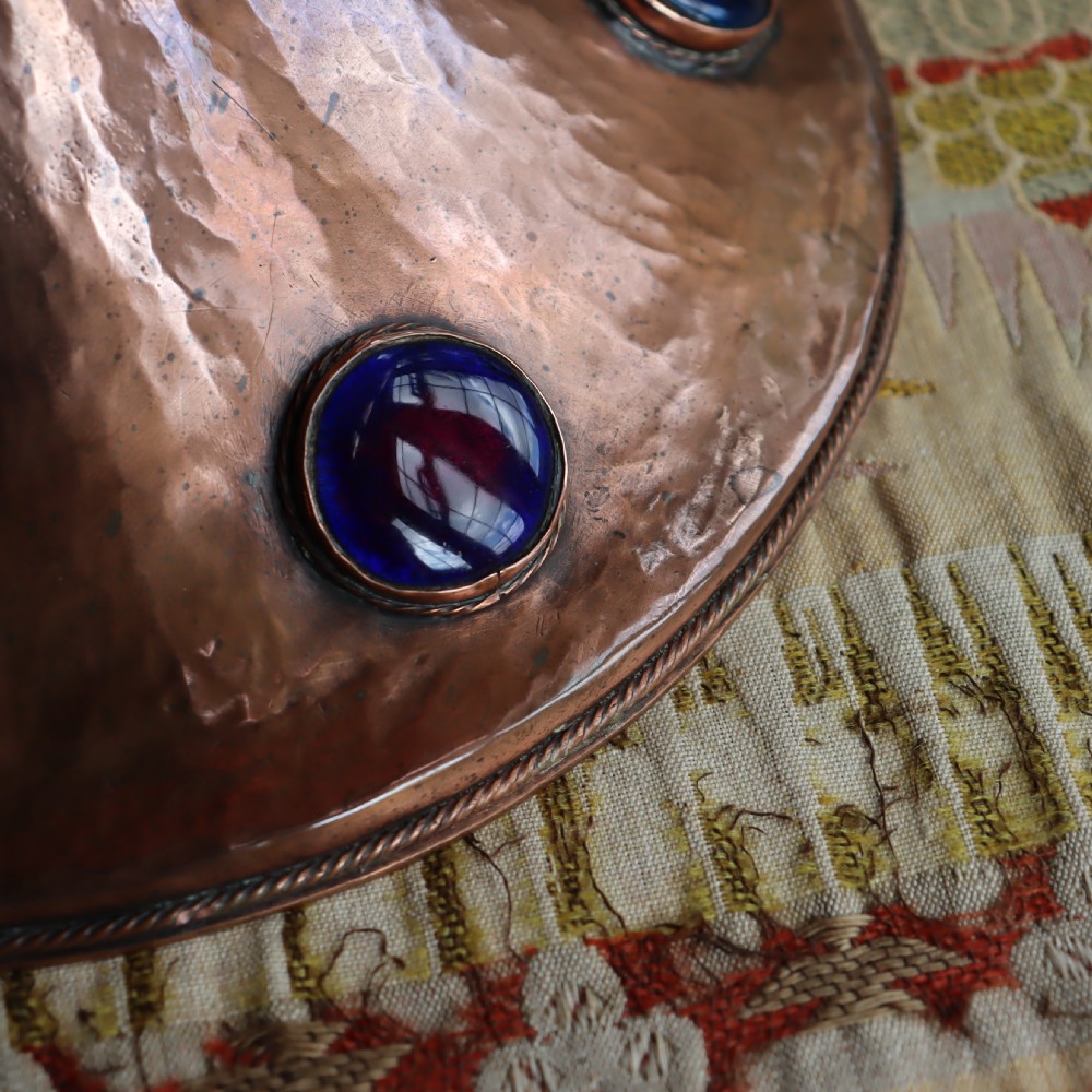 A hand-hammered copper pedestal bowl featuring a flared base set with circular blue enamel cabochons. View V