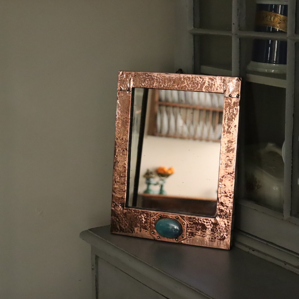 A rectangular hand-hammered copper wall mirror featuring a single blue-green ceramic cabochon mounted at the bottom centre frame.