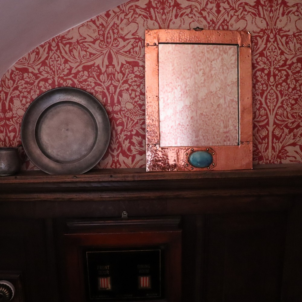 A rectangular hand-hammered copper wall mirror featuring a single blue-green ceramic cabochon mounted at the bottom centre frame. View V