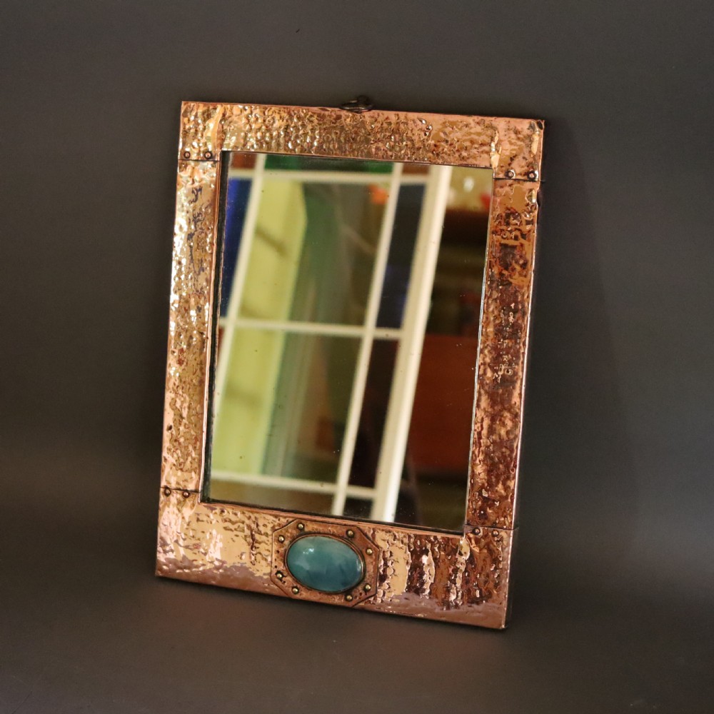 A rectangular hand-hammered copper wall mirror featuring a single blue-green ceramic cabochon mounted at the bottom centre frame. View VI