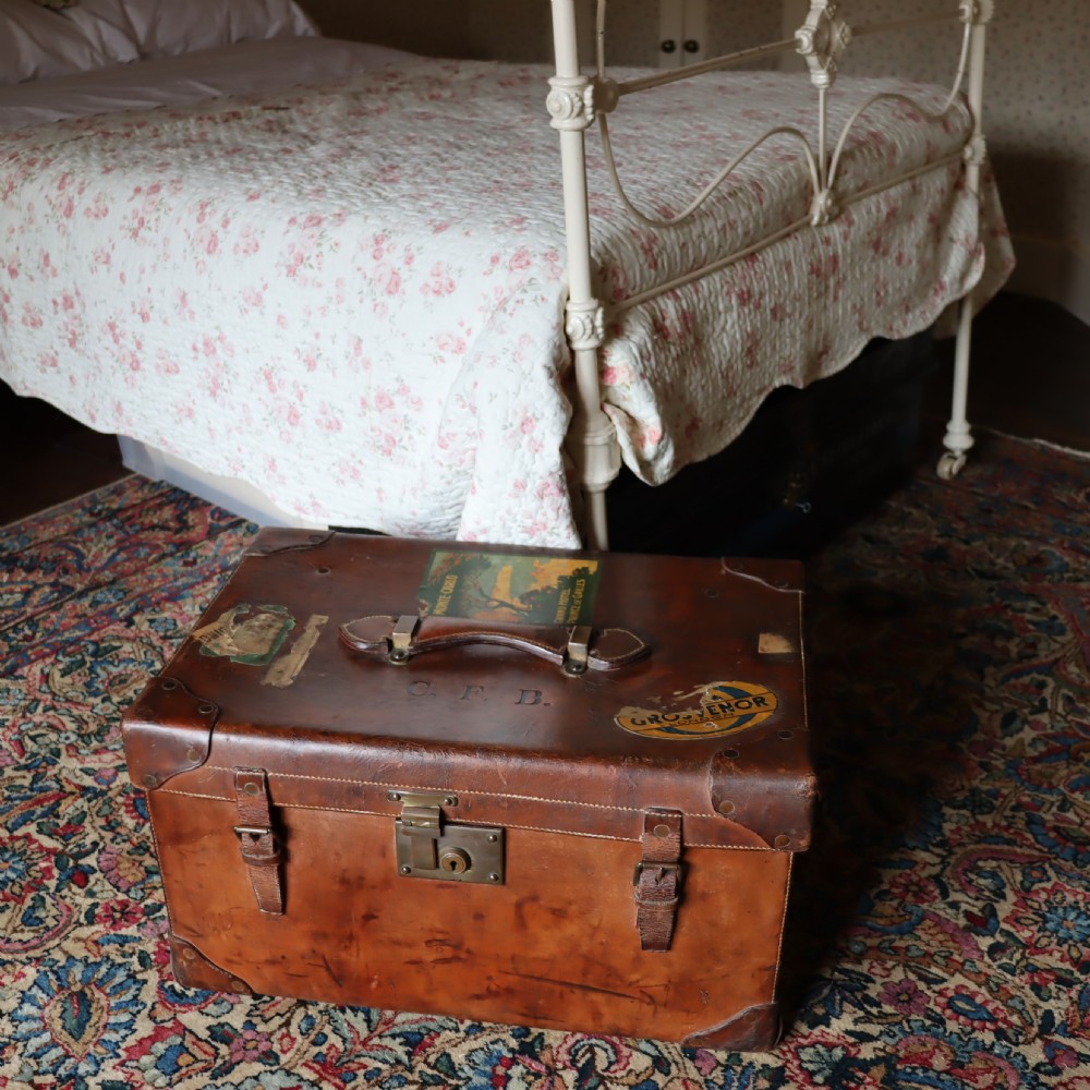 An Edwardian warm brown leather travel trunk with brass locks, leather straps, stencilled monogram initials, and vintage hotel labels.
