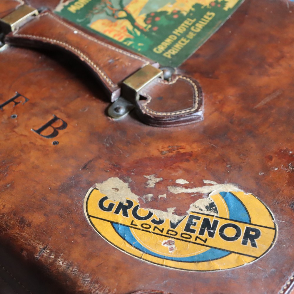 An Edwardian warm brown leather travel trunk with brass locks, leather straps, stencilled monogram initials, and vintage hotel labels. View VI