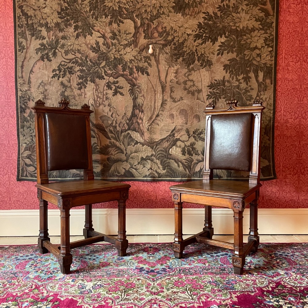 A pair of Victorian Gothic Revival oak hall chairs featuring carved cresting, leather upholstered back panels, solid oak seats, and rectangular stretchers.