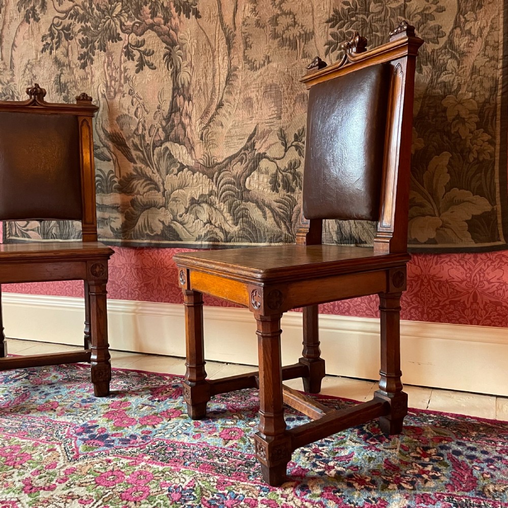 A pair of Victorian Gothic Revival oak hall chairs featuring carved cresting, leather upholstered back panels, solid oak seats, and rectangular stretchers. View II