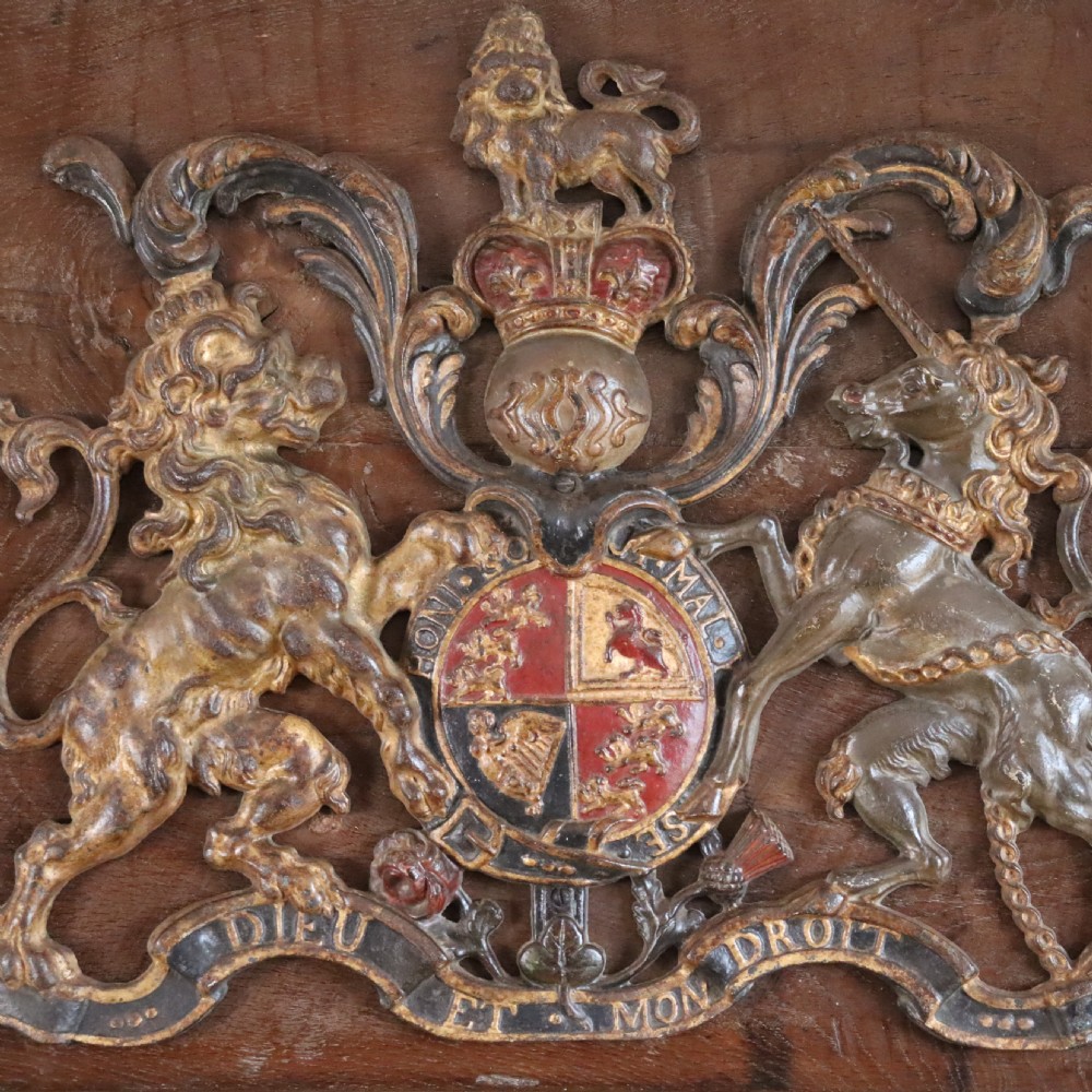 A square dark wood panel featuring a central relief of the British Royal coat of arms with gilded details and metal hanging fixtures at the top. View III