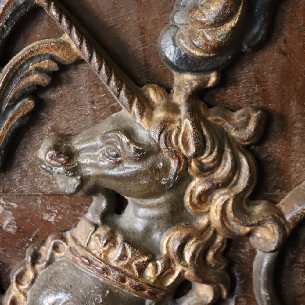 A square dark wood panel featuring a central relief of the British Royal coat of arms with gilded details and metal hanging fixtures at the top. View V