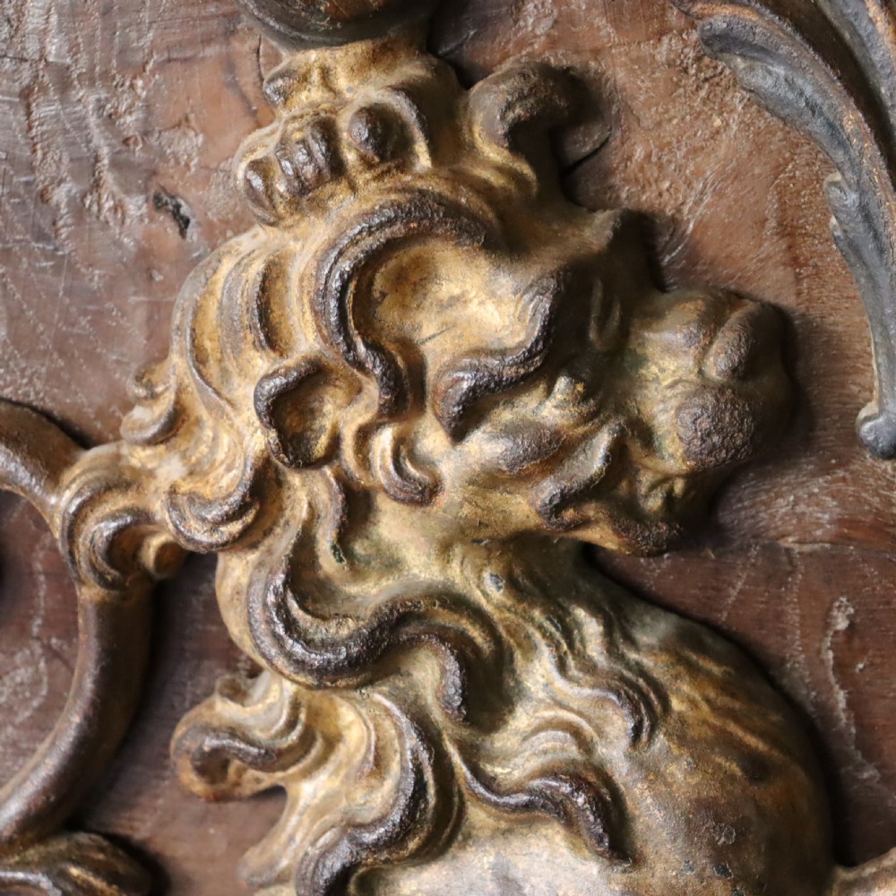 A square dark wood panel featuring a central relief of the British Royal coat of arms with gilded details and metal hanging fixtures at the top. View VI