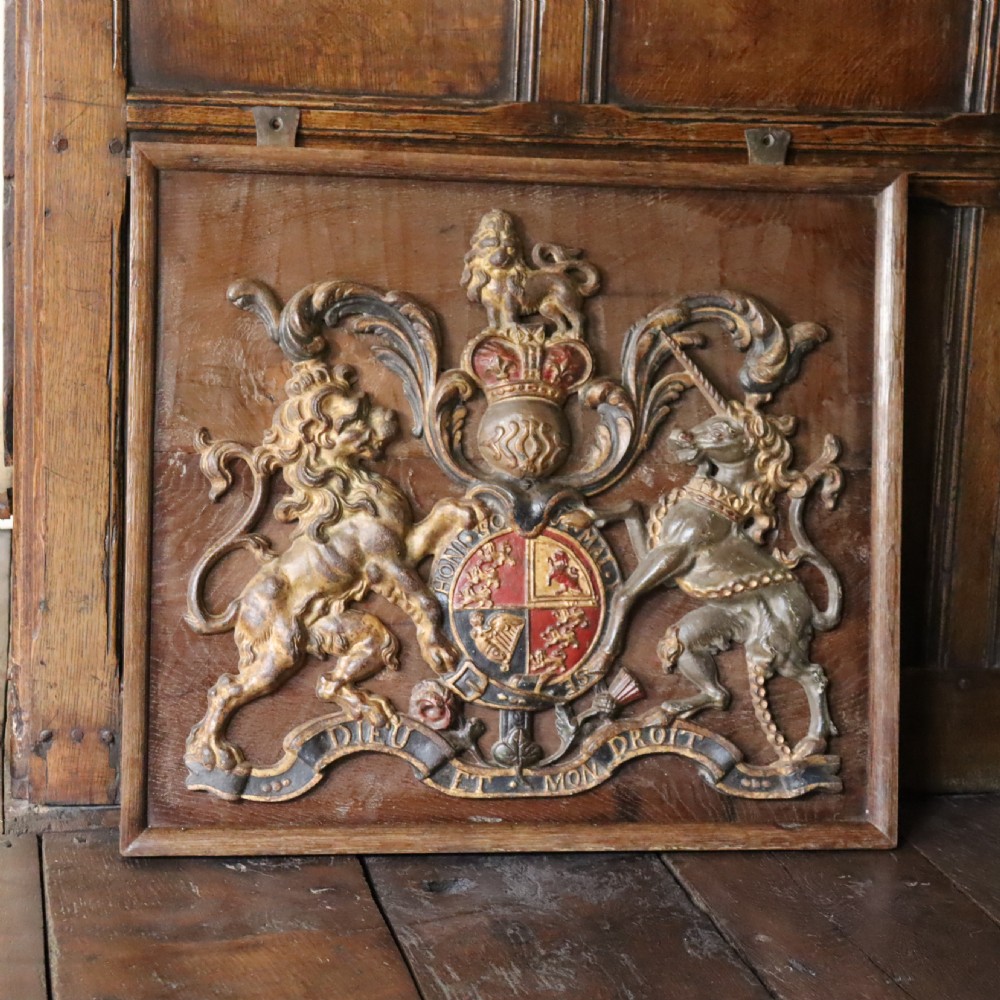 A square dark wood panel featuring a central relief of the British Royal coat of arms with gilded details and metal hanging fixtures at the top. View VII