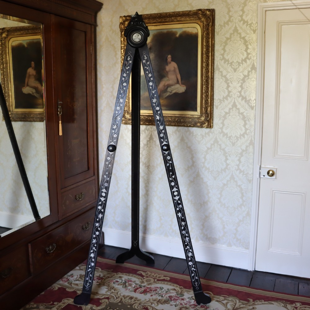 A tall black ebonised gallery easel decorated with white floral inlay along the legs and a carved medallion at the top crest.