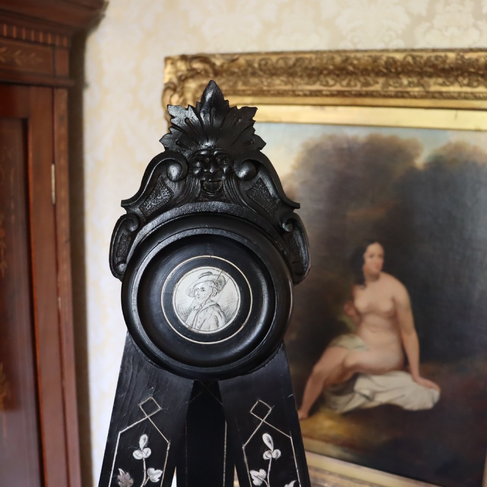 A tall black ebonised gallery easel decorated with white floral inlay along the legs and a carved medallion at the top crest. View V