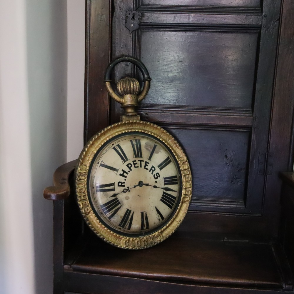 A late 19th-century oversized pocket watch trade sign with a gilded frame, Roman numerals on the dial, and embossed text reading Watches, Jewellery, Clocks. View II