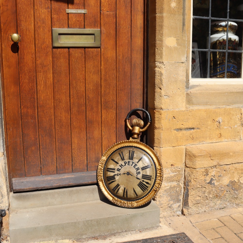 A late 19th-century oversized pocket watch trade sign with a gilded frame, Roman numerals on the dial, and embossed text reading Watches, Jewellery, Clocks. View V