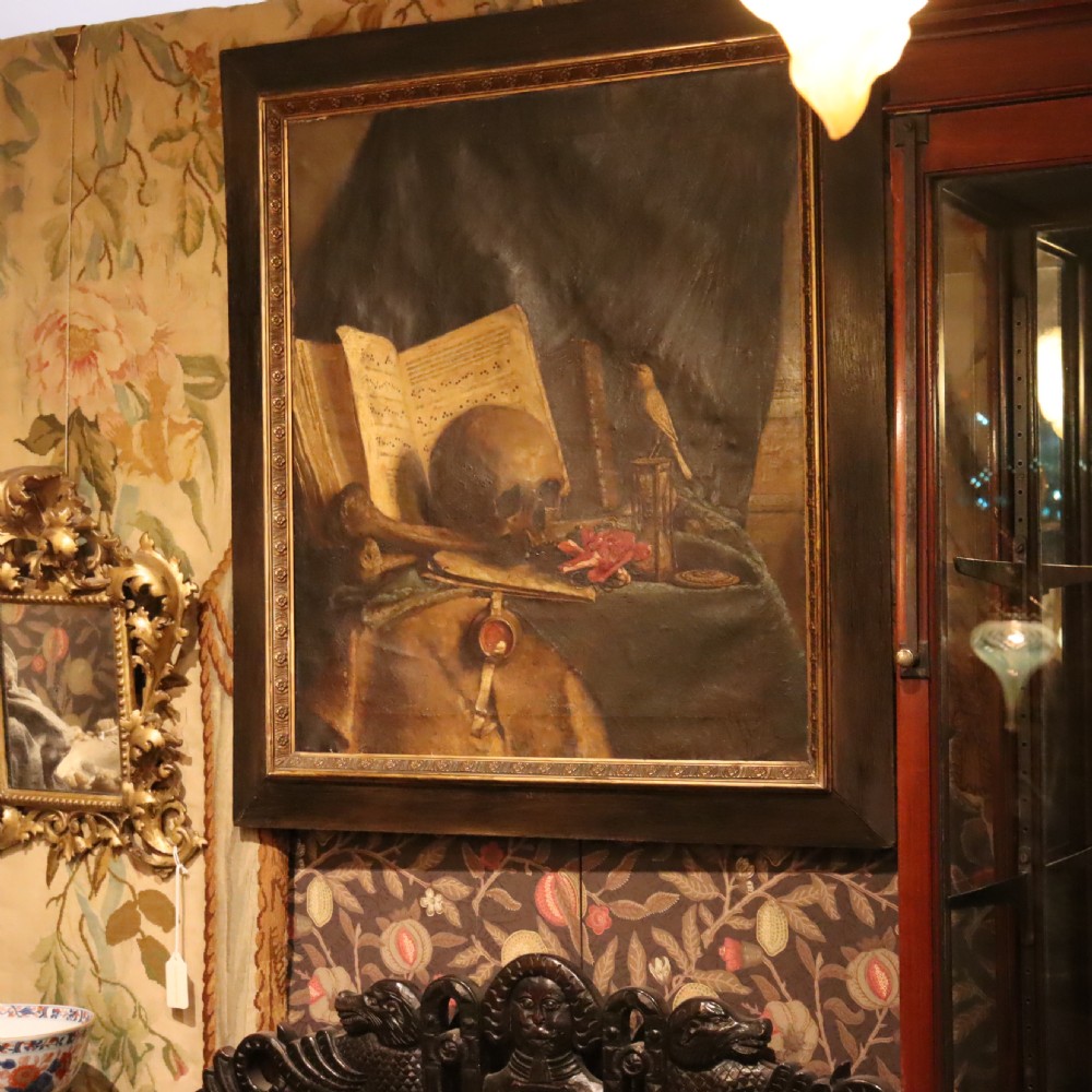 An oil painting depicting a vanitas still life with a human skull, open book, and hourglass set inside a dark moulded frame.
