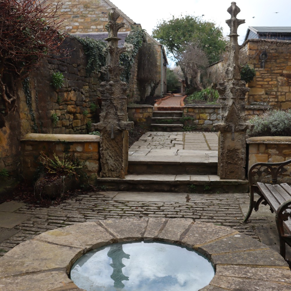 A pair of heavily weathered, hand-carved Gothic limestone pinnacles with intricate crockets and finial tops flanking a stone garden walkway. View III