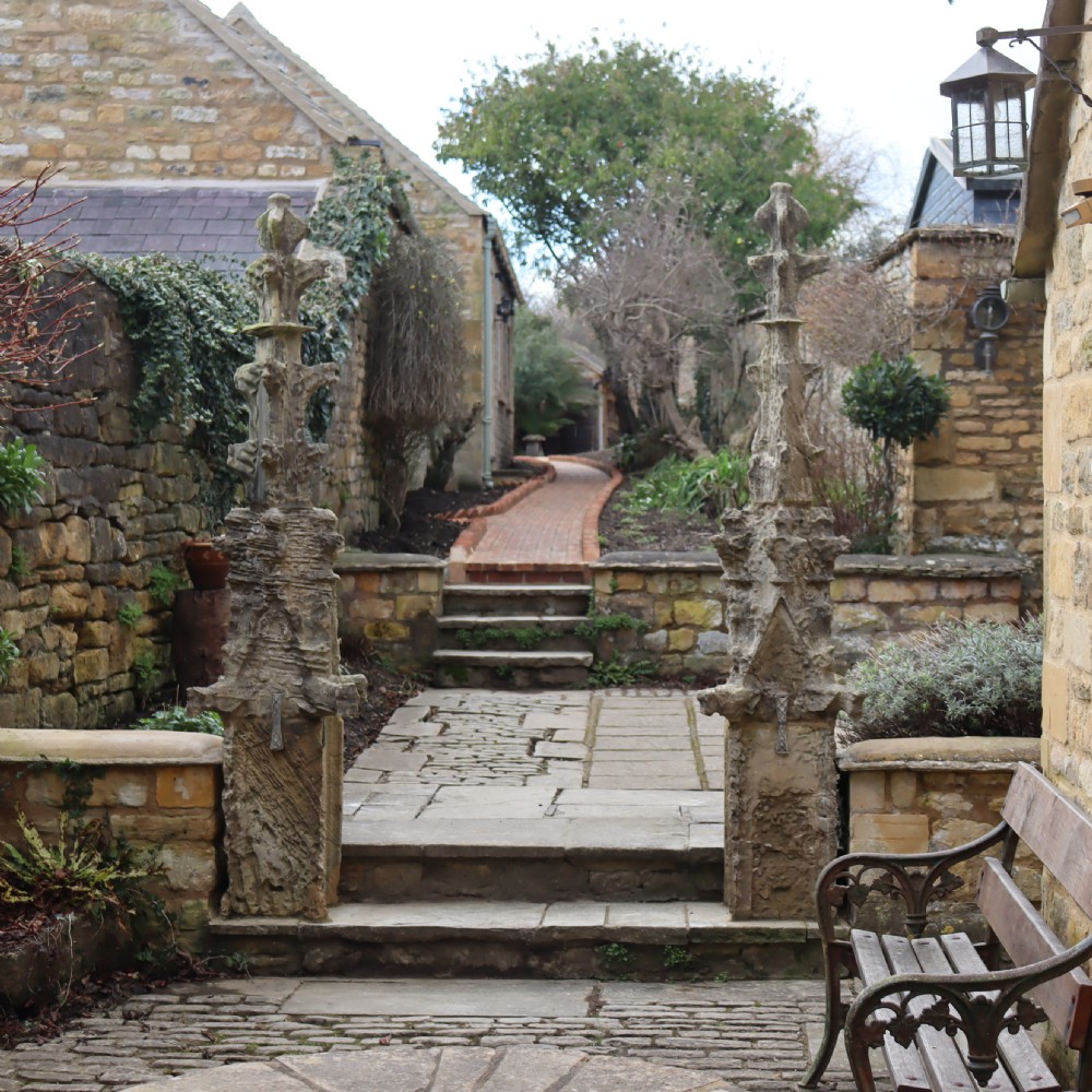 A pair of heavily weathered, hand-carved Gothic limestone pinnacles with intricate crockets and finial tops flanking a stone garden walkway. View II