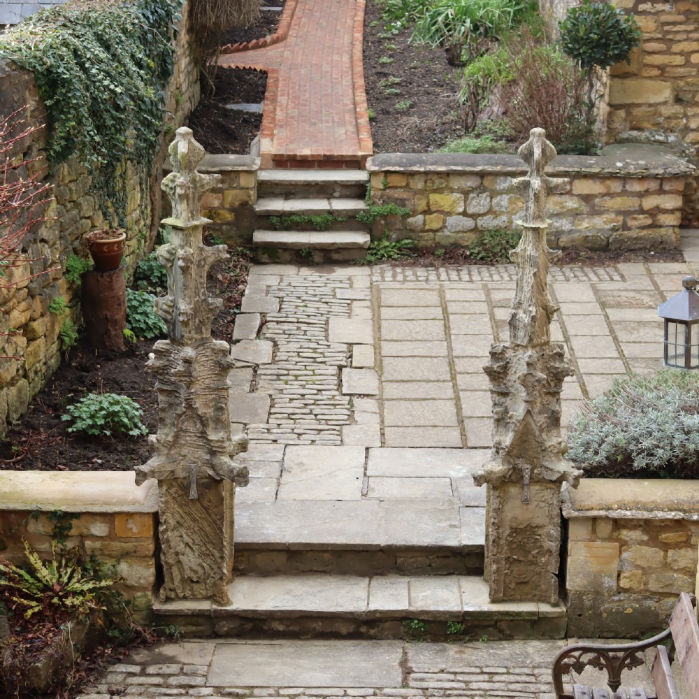 A pair of heavily weathered, hand-carved Gothic limestone pinnacles with intricate crockets and finial tops flanking a stone garden walkway.