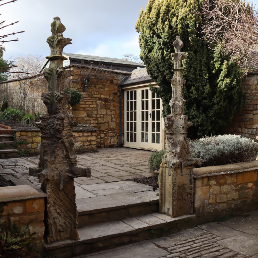 A pair of heavily weathered, hand-carved Gothic limestone pinnacles with intricate crockets and finial tops flanking a stone garden walkway. View IV