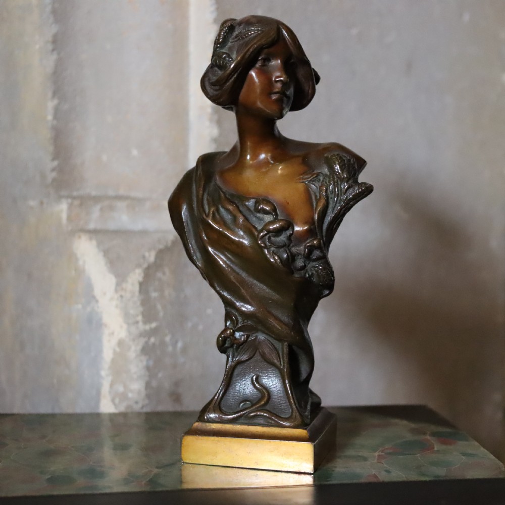 An Art Nouveau patinated bronze bust depicting a young woman adorned with ears of wheat and flowing drapery, set on a gilt base.