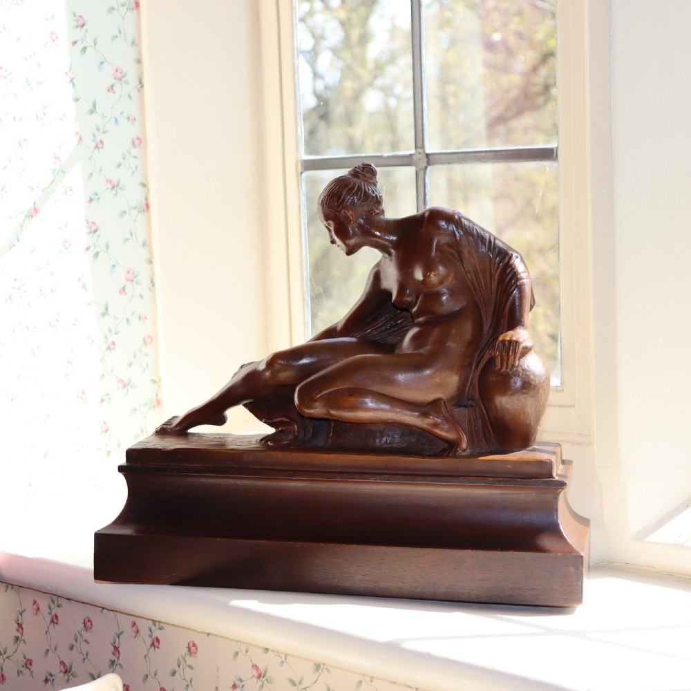 A carved warm brown limewood sculpture depicting a seated female nude figure on a molded rectangular plinth.