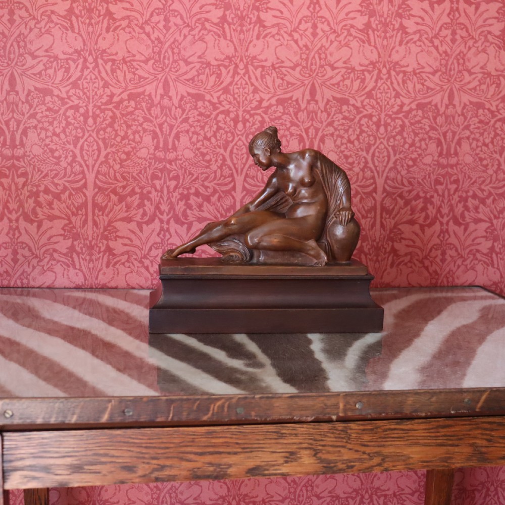 A carved warm brown limewood sculpture depicting a seated female nude figure on a molded rectangular plinth. View II