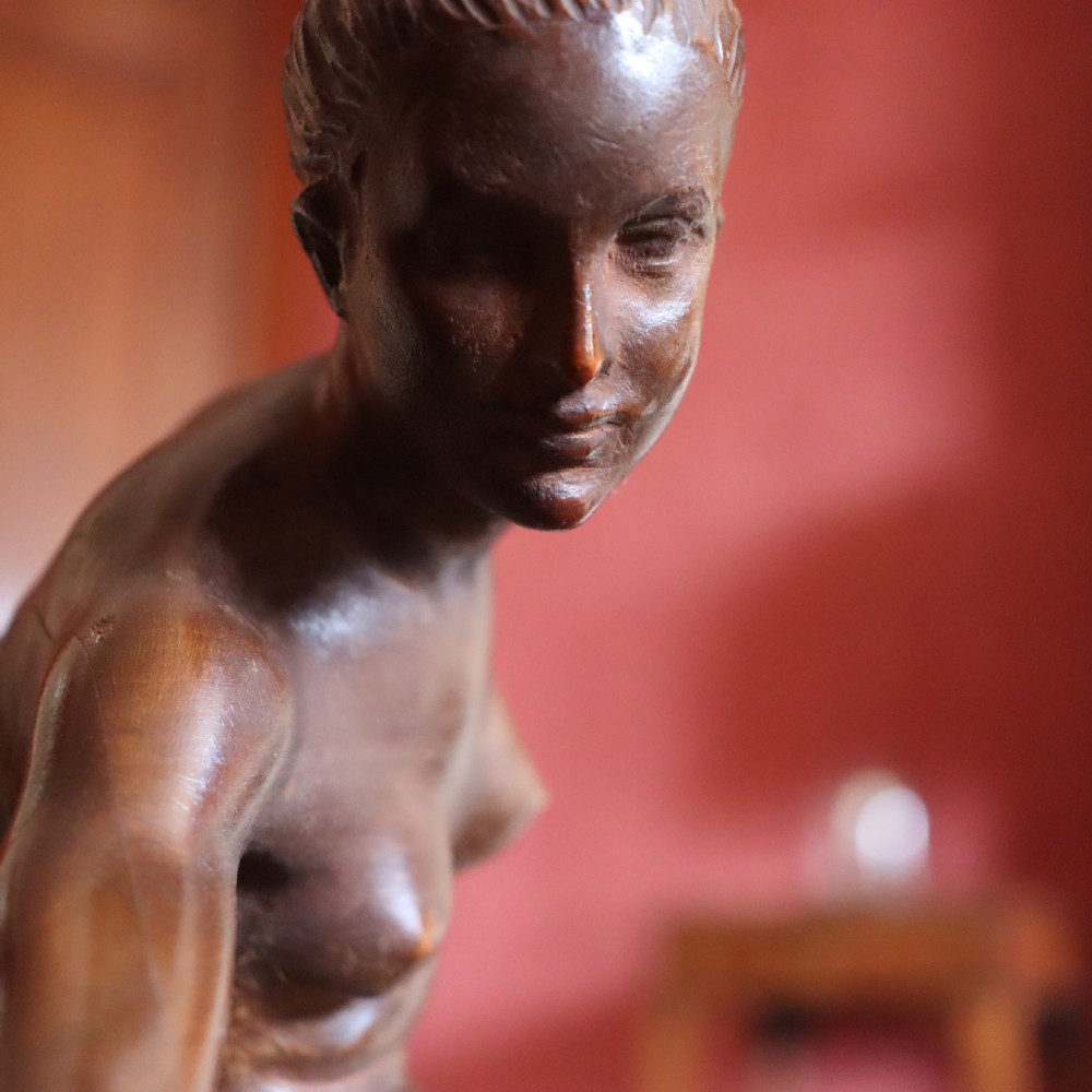 A carved warm brown limewood sculpture depicting a seated female nude figure on a molded rectangular plinth. View III