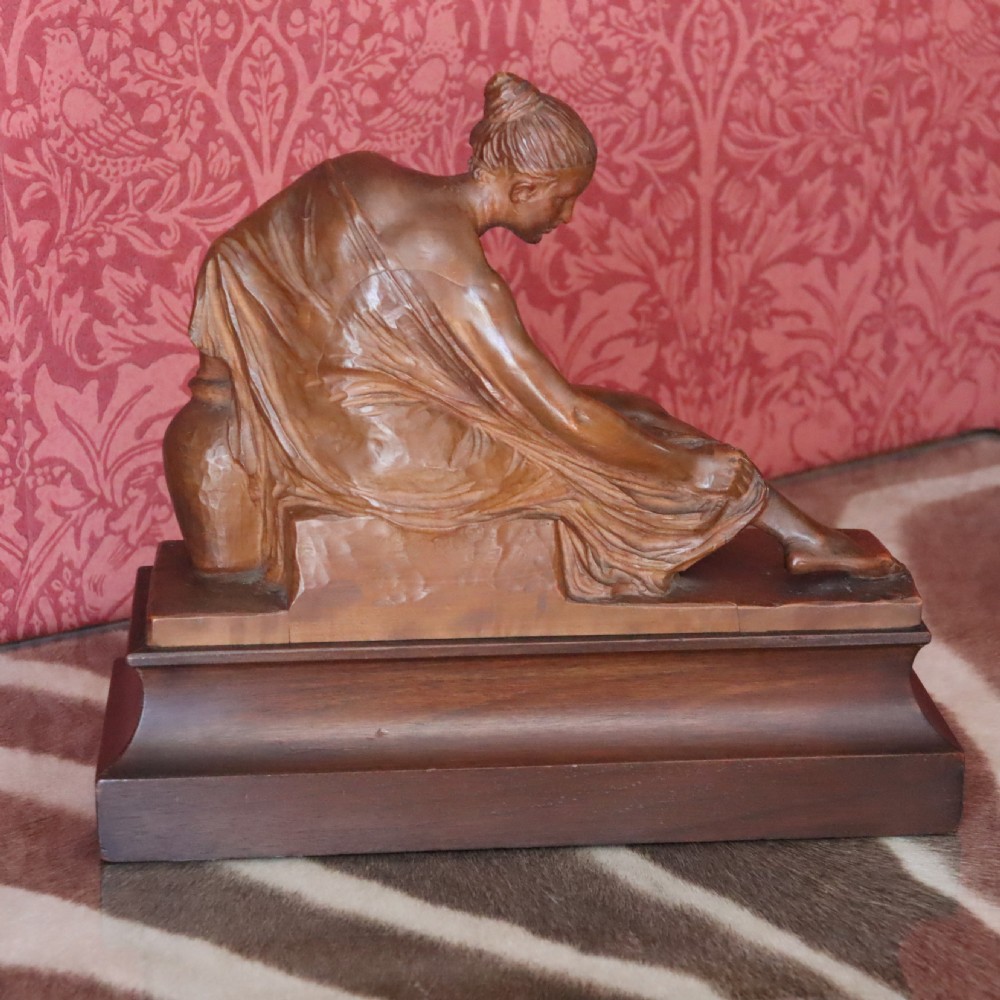 A carved warm brown limewood sculpture depicting a seated female nude figure on a molded rectangular plinth. View IV
