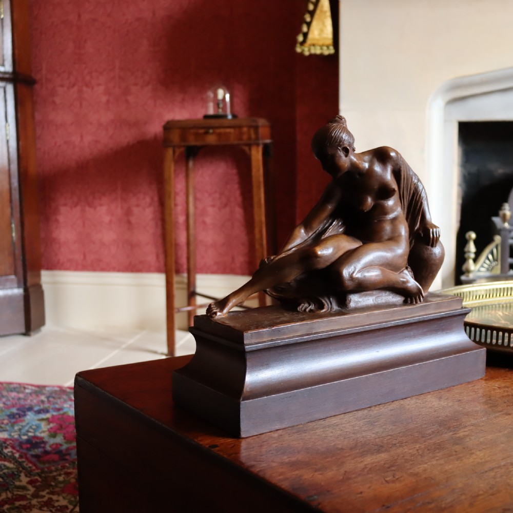 A carved warm brown limewood sculpture depicting a seated female nude figure on a molded rectangular plinth. View VIII