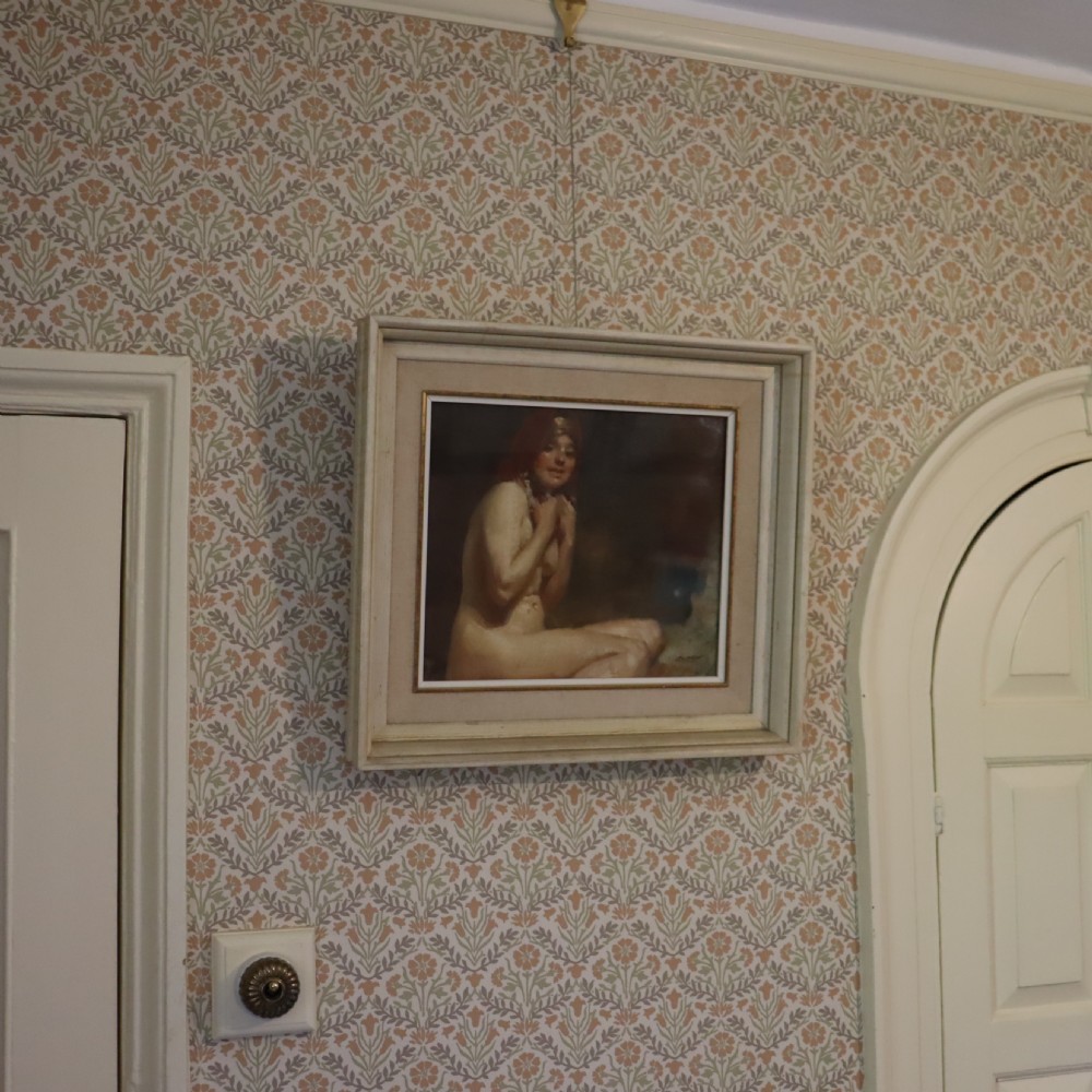 A framed antique oil painting depicting a seated female nude with a red head covering, presented in a light cream moulded wooden frame. View VIII