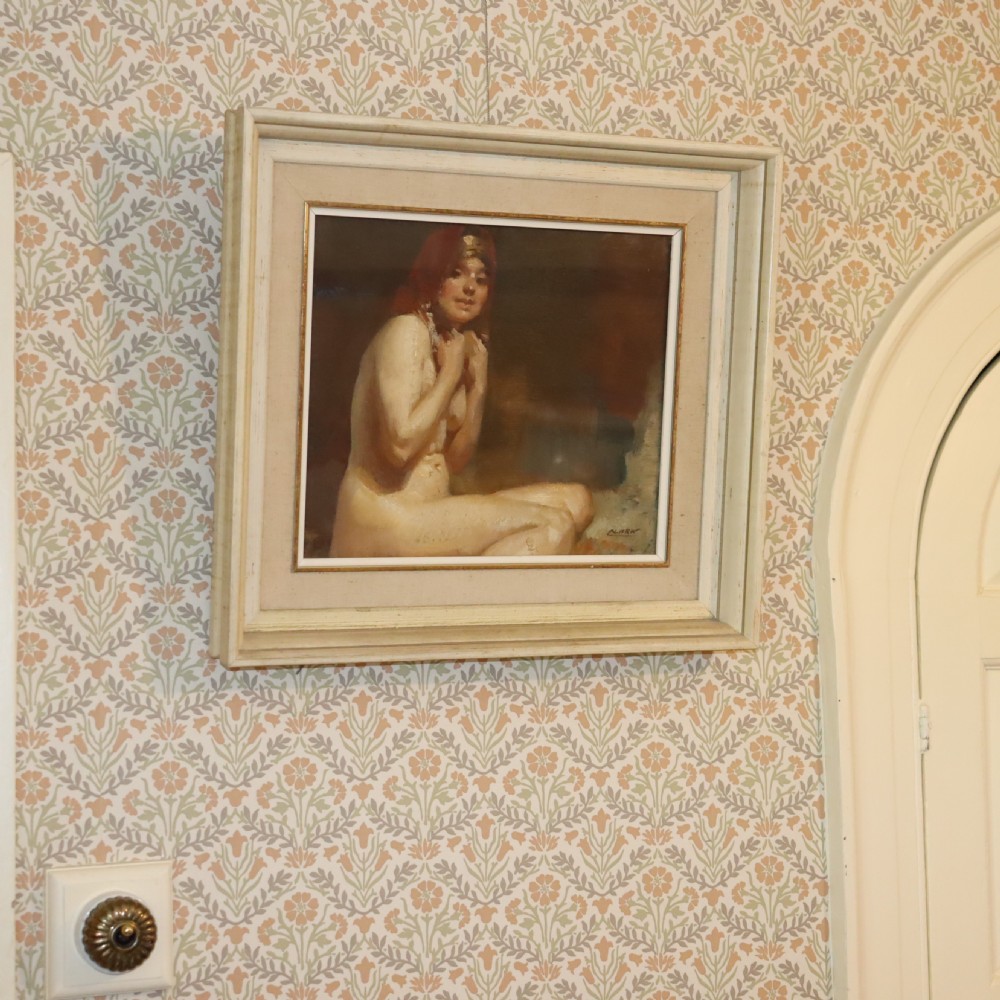 A framed antique oil painting depicting a seated female nude with a red head covering, presented in a light cream moulded wooden frame.