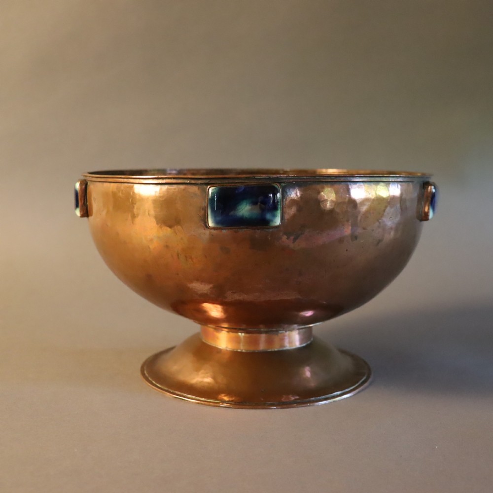 A hand-hammered copper pedestal bowl featuring rectangular blue-glazed ceramic cabochons mounted around the rim.
