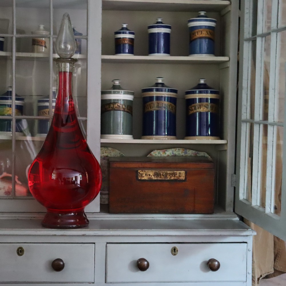 A large teardrop-shaped clear glass apothecary carboy filled with red liquid, featuring a tall slender neck and a glass stopper. View IV