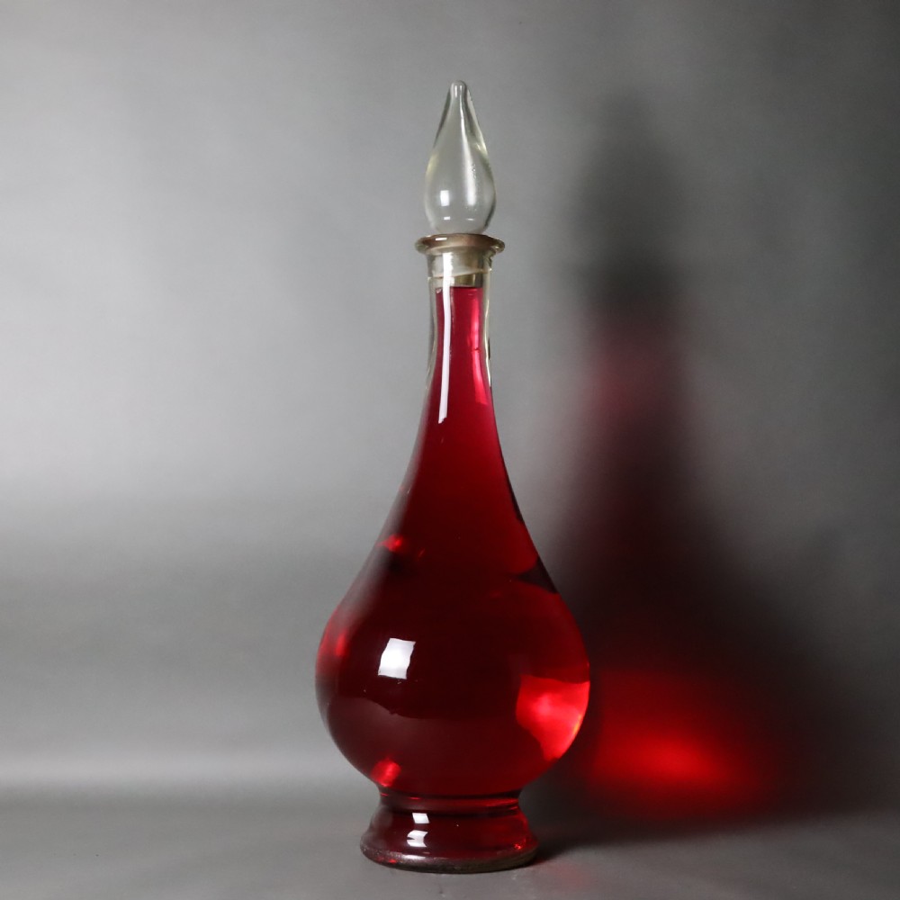 A large teardrop-shaped clear glass apothecary carboy filled with red liquid, featuring a tall slender neck and a glass stopper. View II
