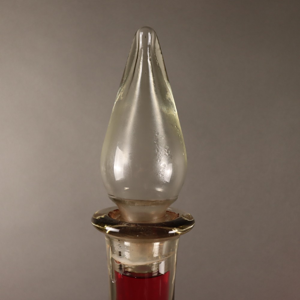 A large teardrop-shaped clear glass apothecary carboy filled with red liquid, featuring a tall slender neck and a glass stopper. View III