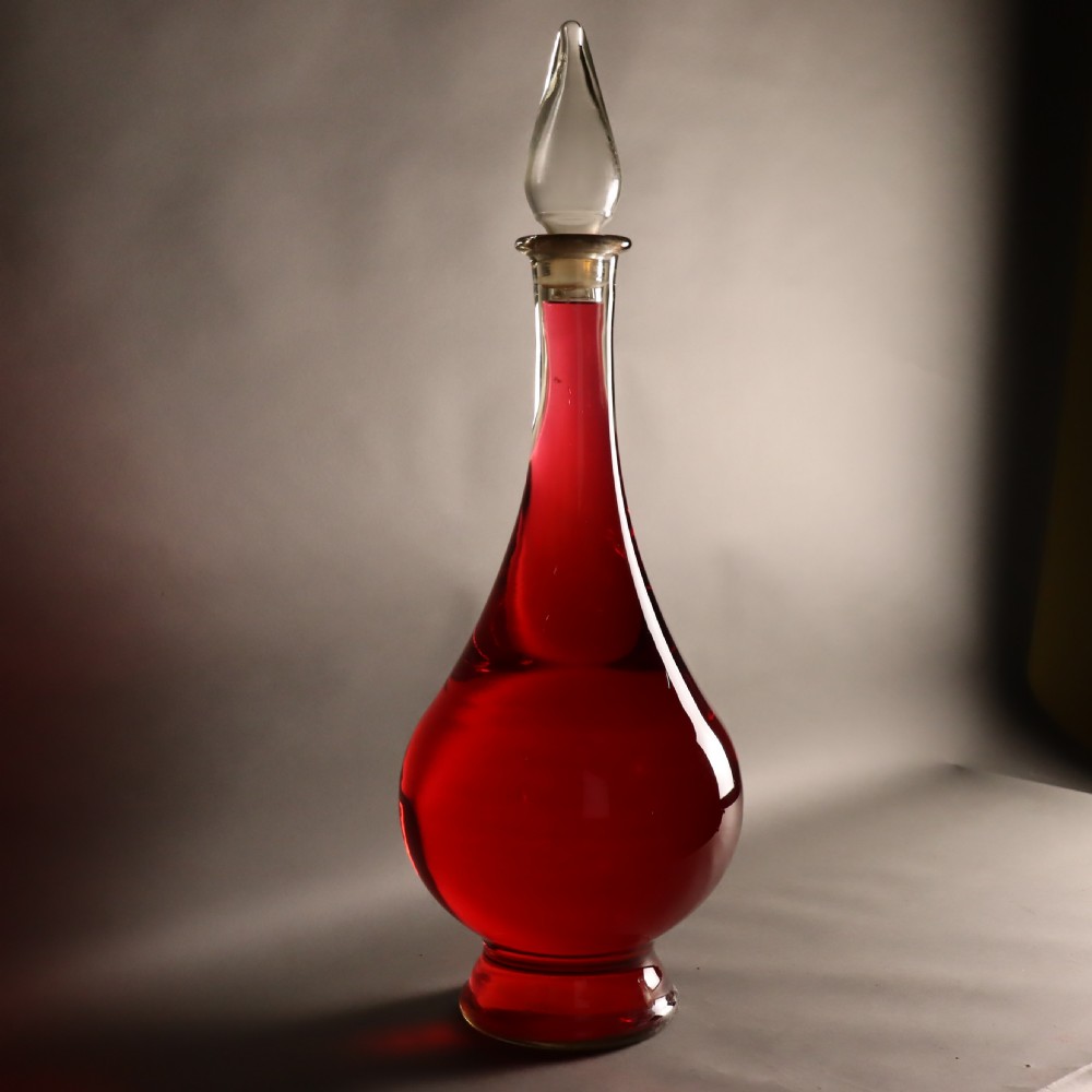 A large teardrop-shaped clear glass apothecary carboy filled with red liquid, featuring a tall slender neck and a glass stopper.